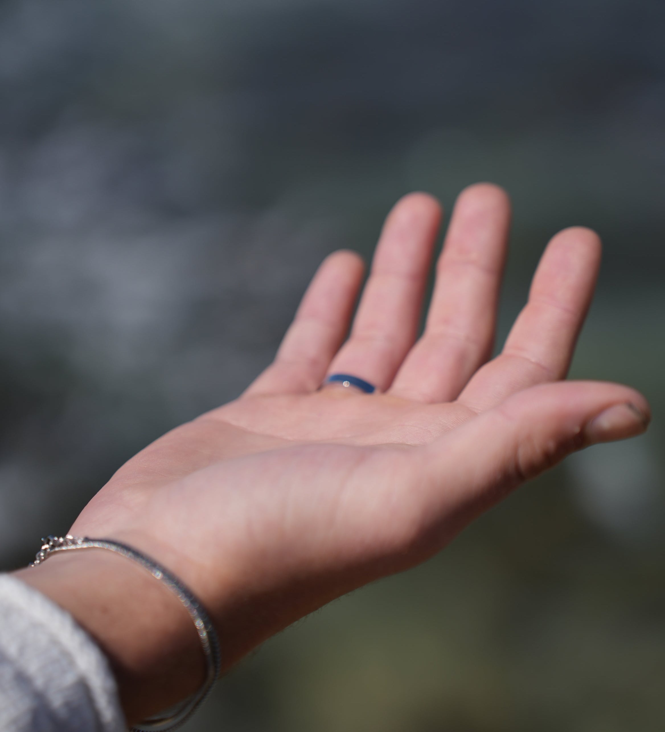 תקריב של יד גבר עונדת צמיד כסף עדין וטבעת עם אבן כחולה על רקע ים מטושטש, להמחשת תכשיט לגבר עם משמעות.