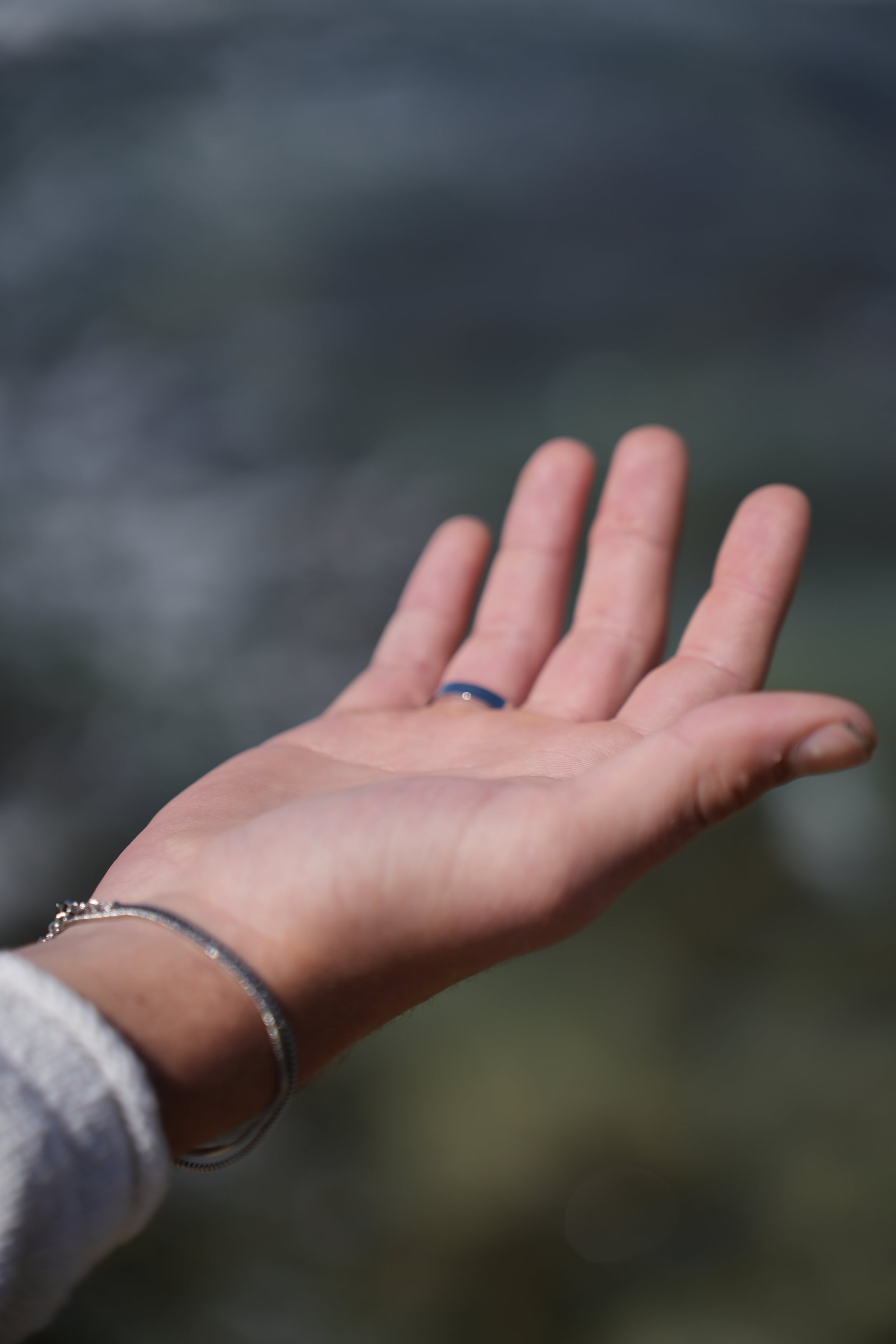 תקריב של יד גבר עונדת צמיד כסף עדין וטבעת עם אבן כחולה על רקע ים מטושטש, להמחשת תכשיט לגבר עם משמעות.