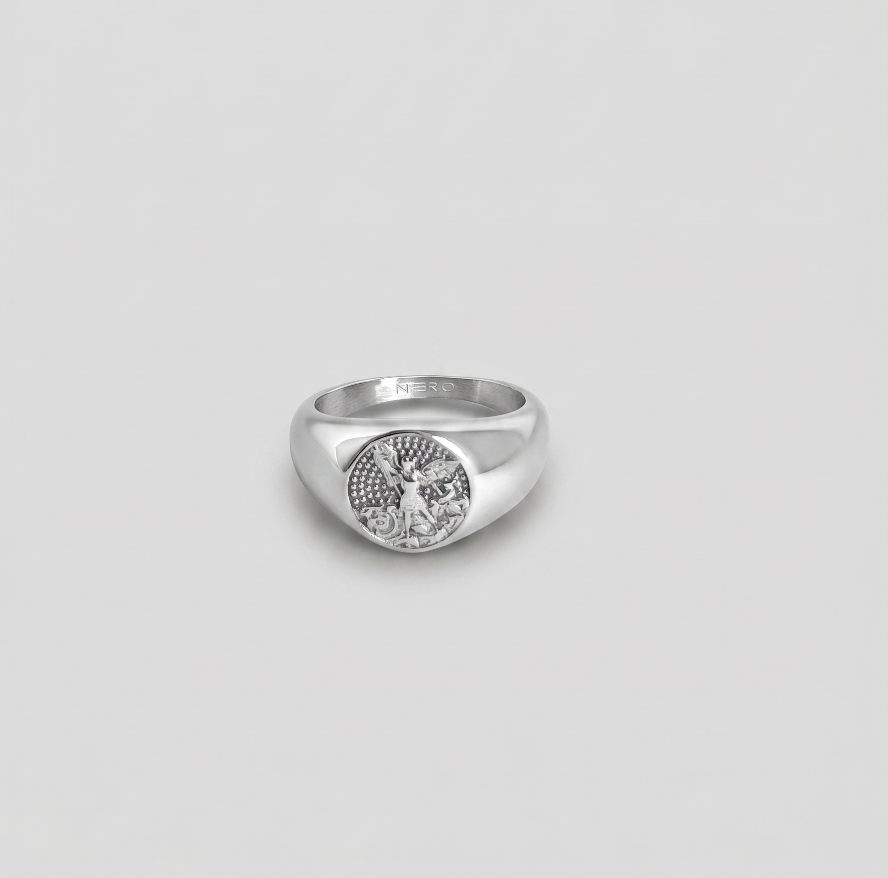 טבעת חותם (Signet ring) גברית בגוון כסף מבריק, עשויה פלדת אל-חלד. על החותם המעוגל חרוטה דמות שומר מפורטת בסגנון קלאסי על רקע טקסטורת נקודות. על הדופן הפנימית של הטבעת חרוט המותג "NERO".
