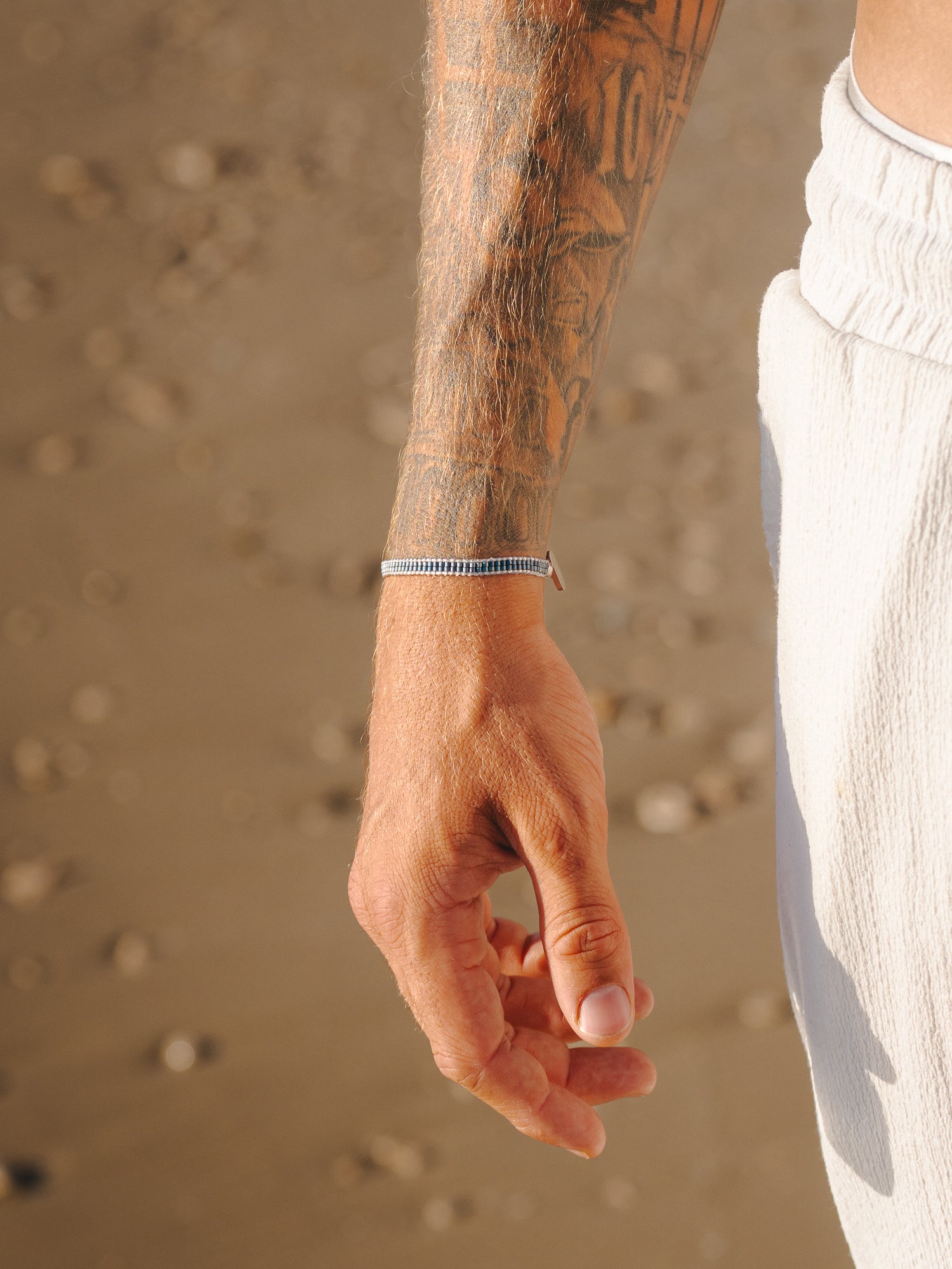 צמיד Aqua כחול כהה על פרק כף יד מקועקעת של גבר, על רקע חול בחוף.