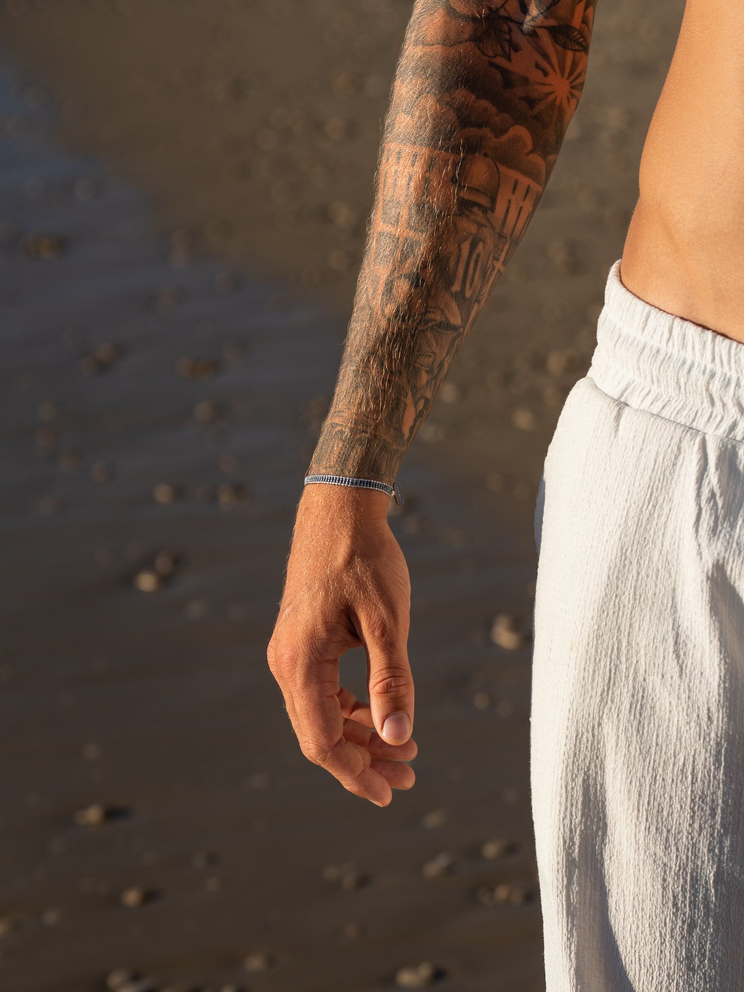 צמיד Aqua כחול כהה על פרק כף יד מקועקעת, על רקע חוף ים או חול.