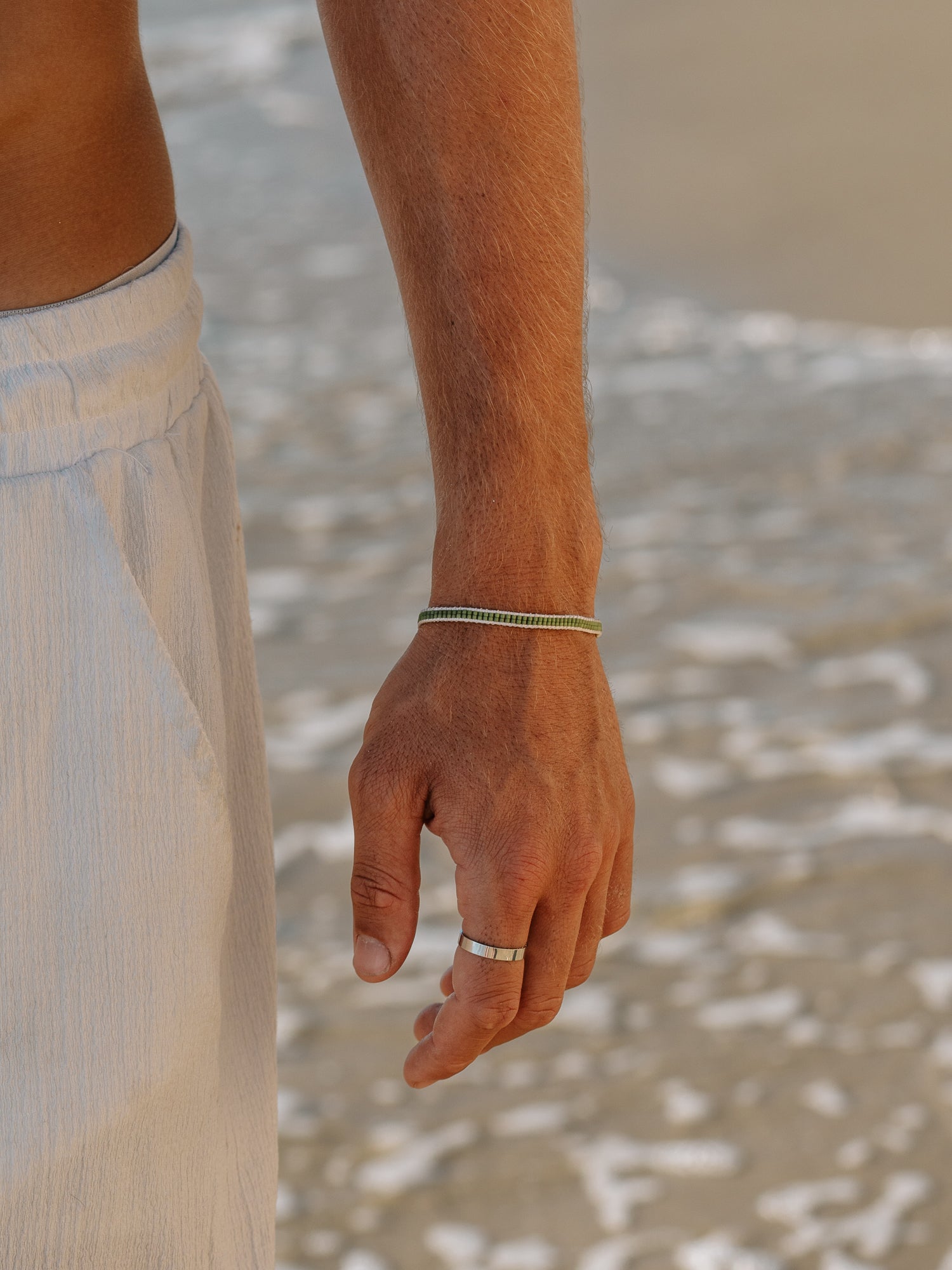 צמיד Aqua ירוק זית על פרק כף יד של גבר, על רקע מים גולשים וחול בחוף, טבעת כסף על האצבע.