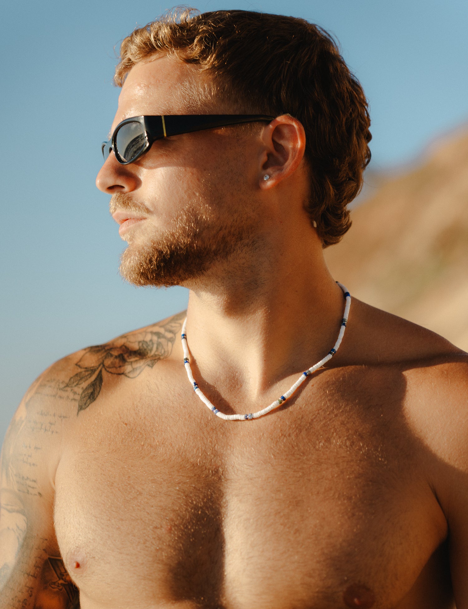 גבר חזה חשוף בחוף, לובש משקפי שמש ושרשרת Arctic לבן-כחול.