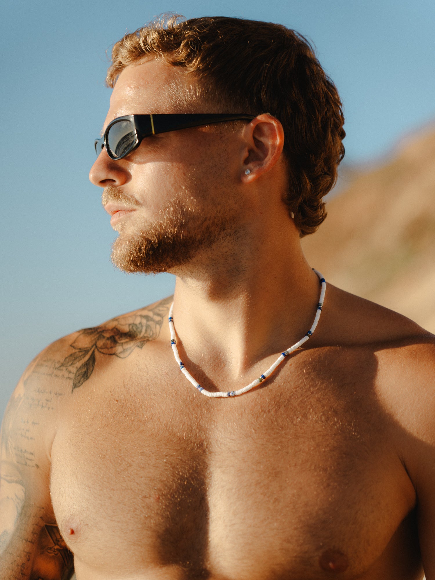 גבר חזה חשוף בחוף, לובש משקפי שמש ושרשרת Arctic לבן-כחול.