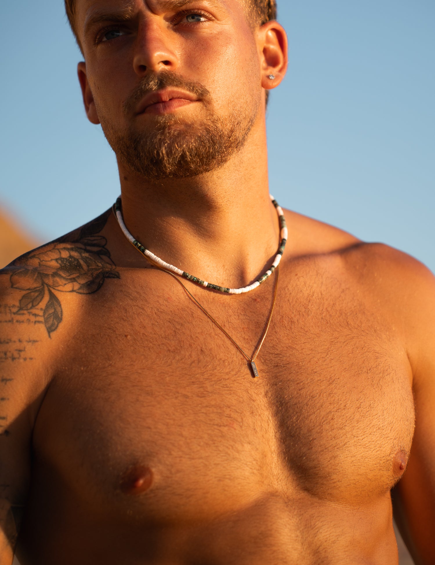 תקריב על פניו וחזהו של גבר ללא חולצה בחוף, לובש שרשרת Azure ושכבה נוספת של שרשרת דקה.