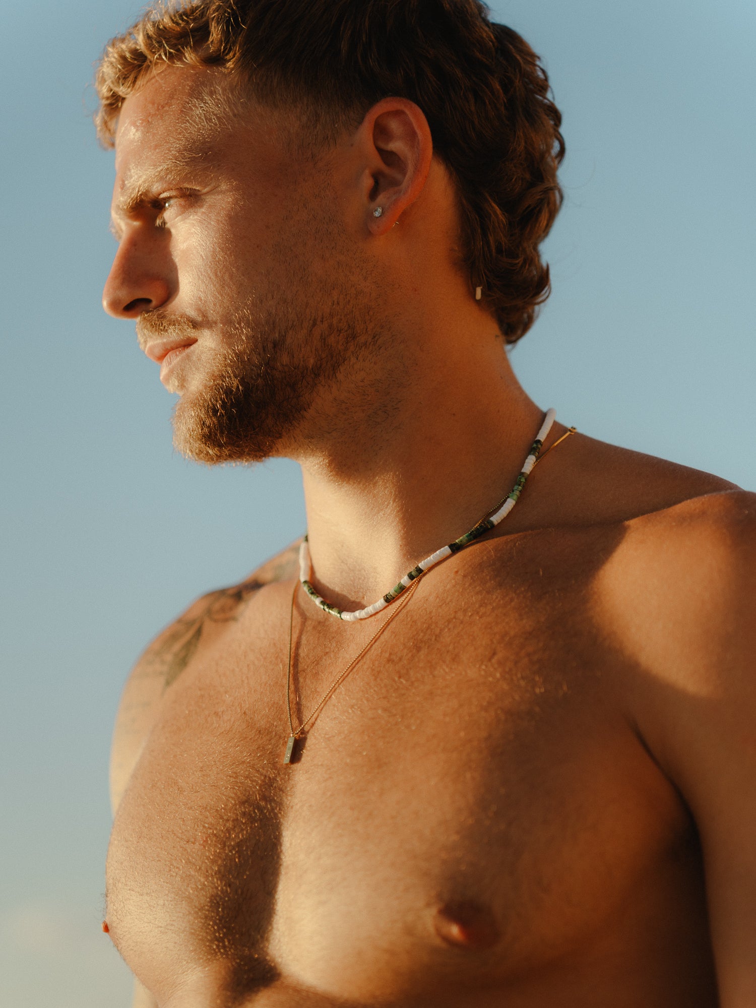 גבר חזה חשוף לובש שתי שרשראות בחוף: שרשרת Azure מחרוזים ואבן תליון קטנה, ושכבה של שרשרת דקה יותר עם תליון מלבני.