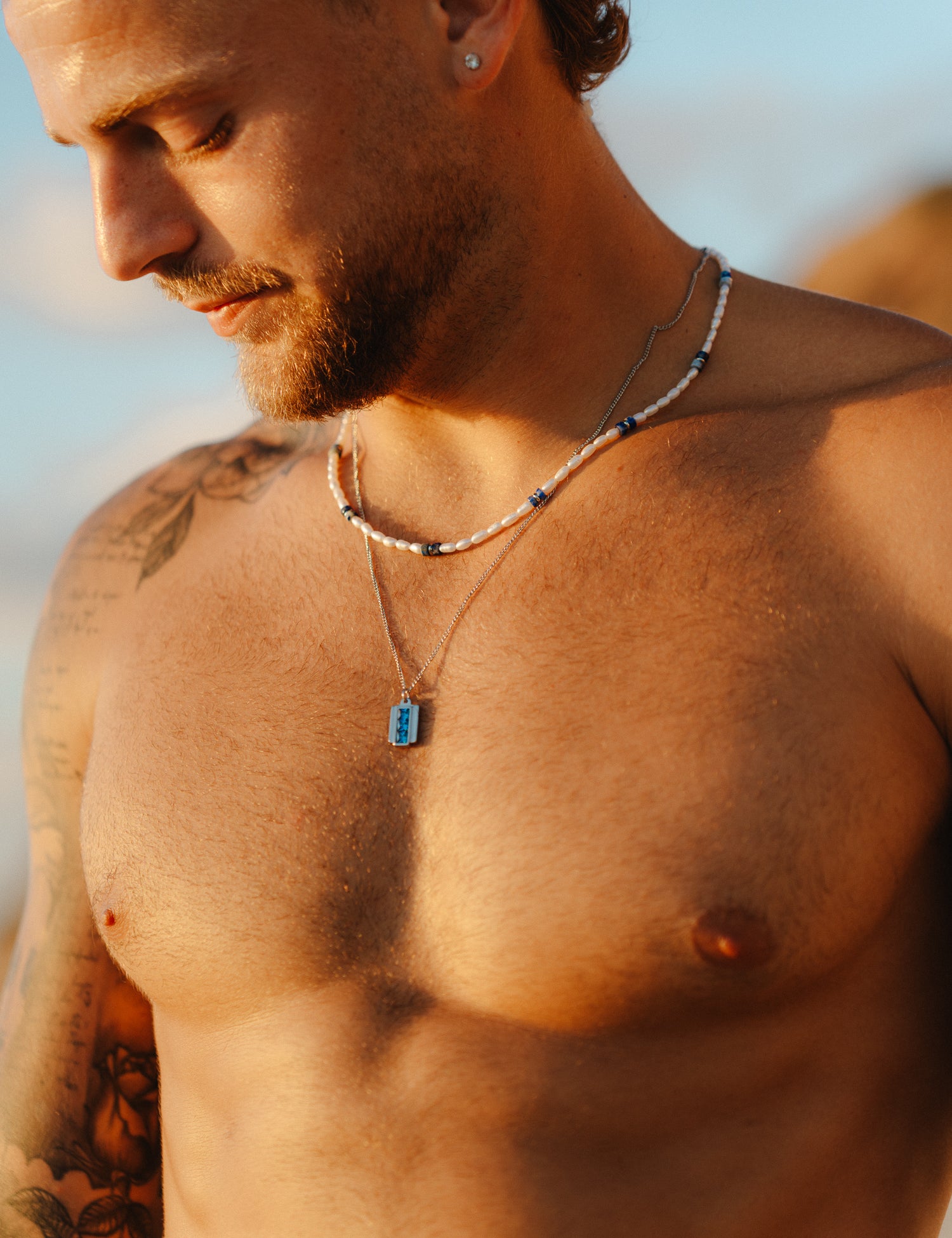 גבר חזה חשוף לובש את שרשרת Beluga מפנינים ולפיס לזולי בחוף, בשילוב שתי שרשראות כסף נוספות.