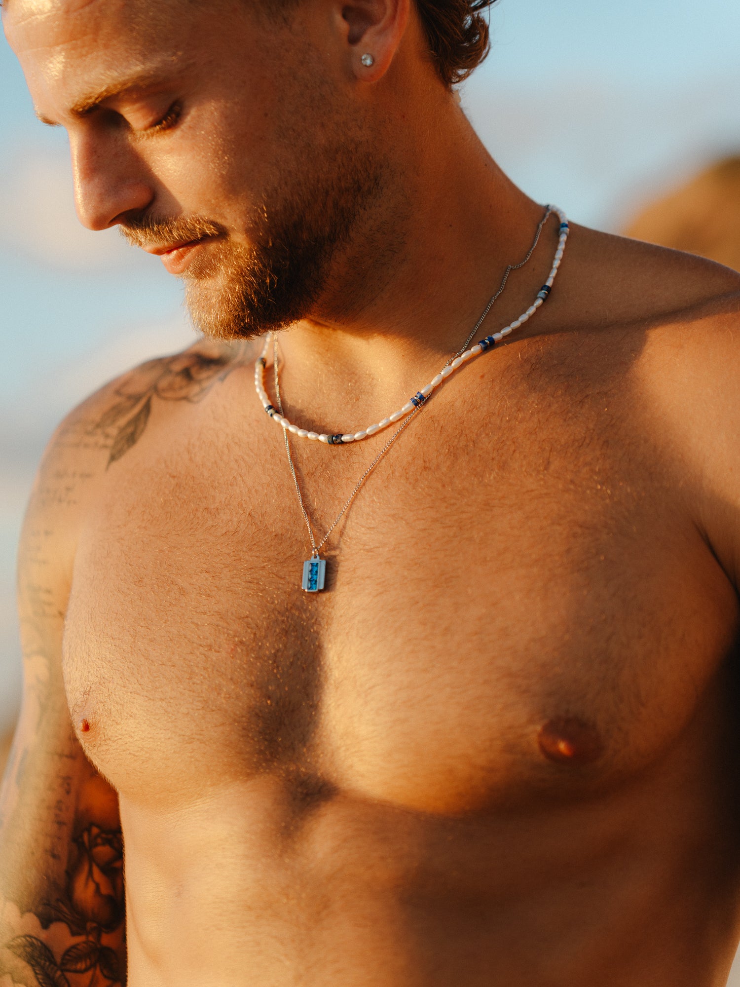 גבר חזה חשוף לובש את שרשרת Beluga מפנינים ולפיס לזולי בחוף, בשילוב שתי שרשראות כסף נוספות.