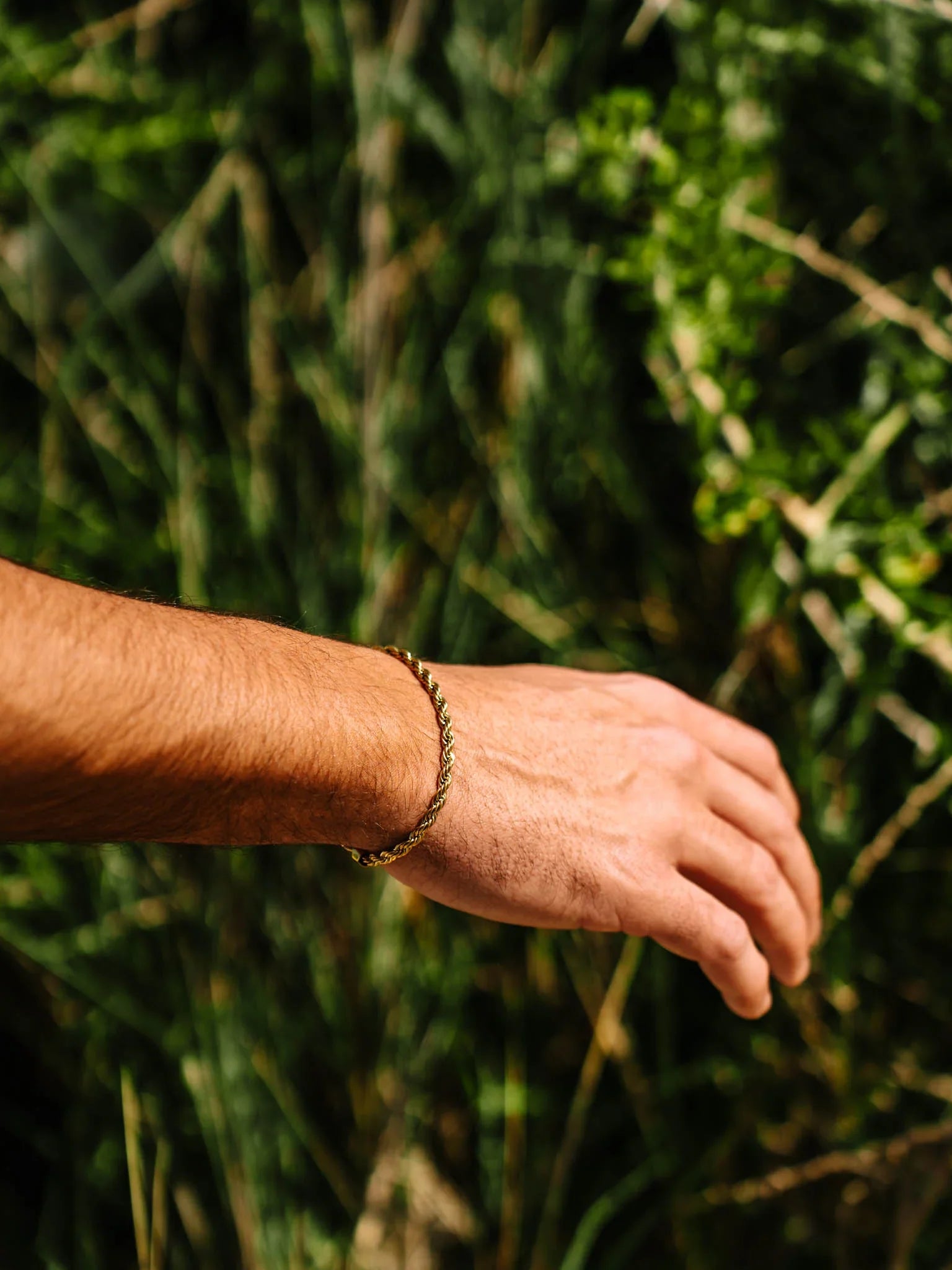 יד של גבר עם צמיד Eros מוזהב על פרק כף היד, מצולם באור שמש טבעי מול רקע של צמחייה ירוקה.