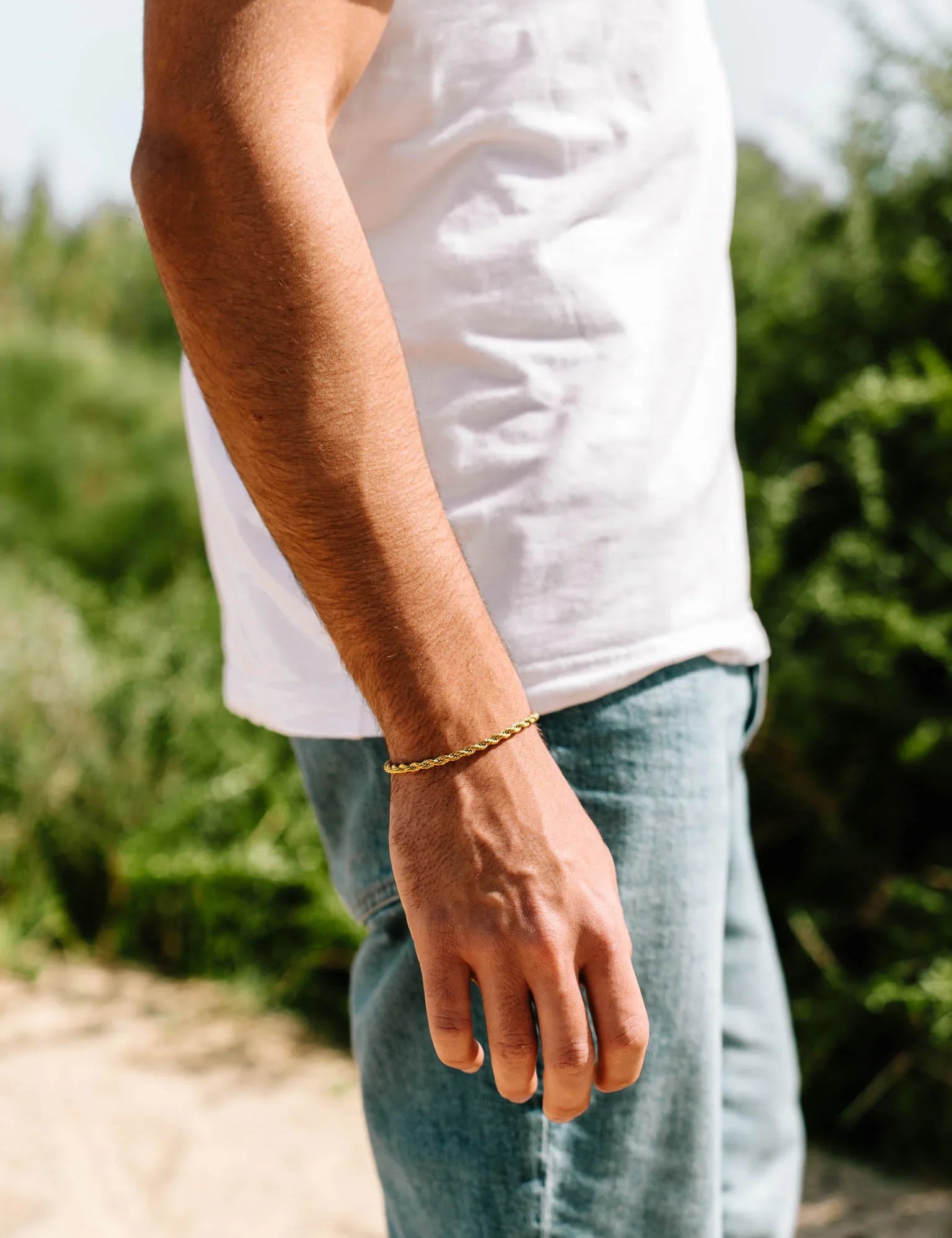 יד של גבר עם צמיד Eros מוזהב על פרק כף היד, מצולם באור טבעי ליד צמחייה, לבוש חולצה לבנה ומכנסי ג'ינס.