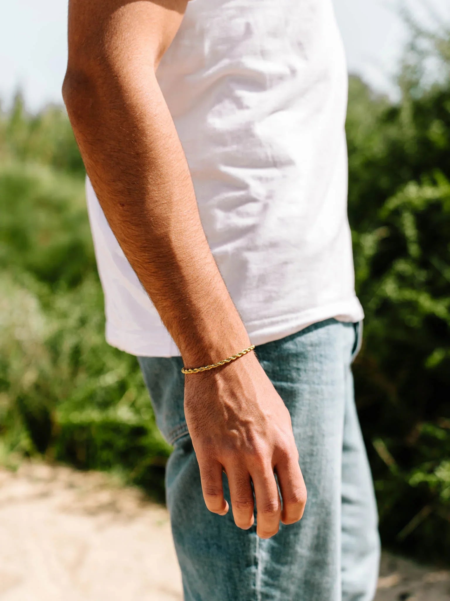 יד של גבר עם צמיד Eros מוזהב על פרק כף היד, מצולם באור טבעי ליד צמחייה, לבוש חולצה לבנה ומכנסי ג'ינס.