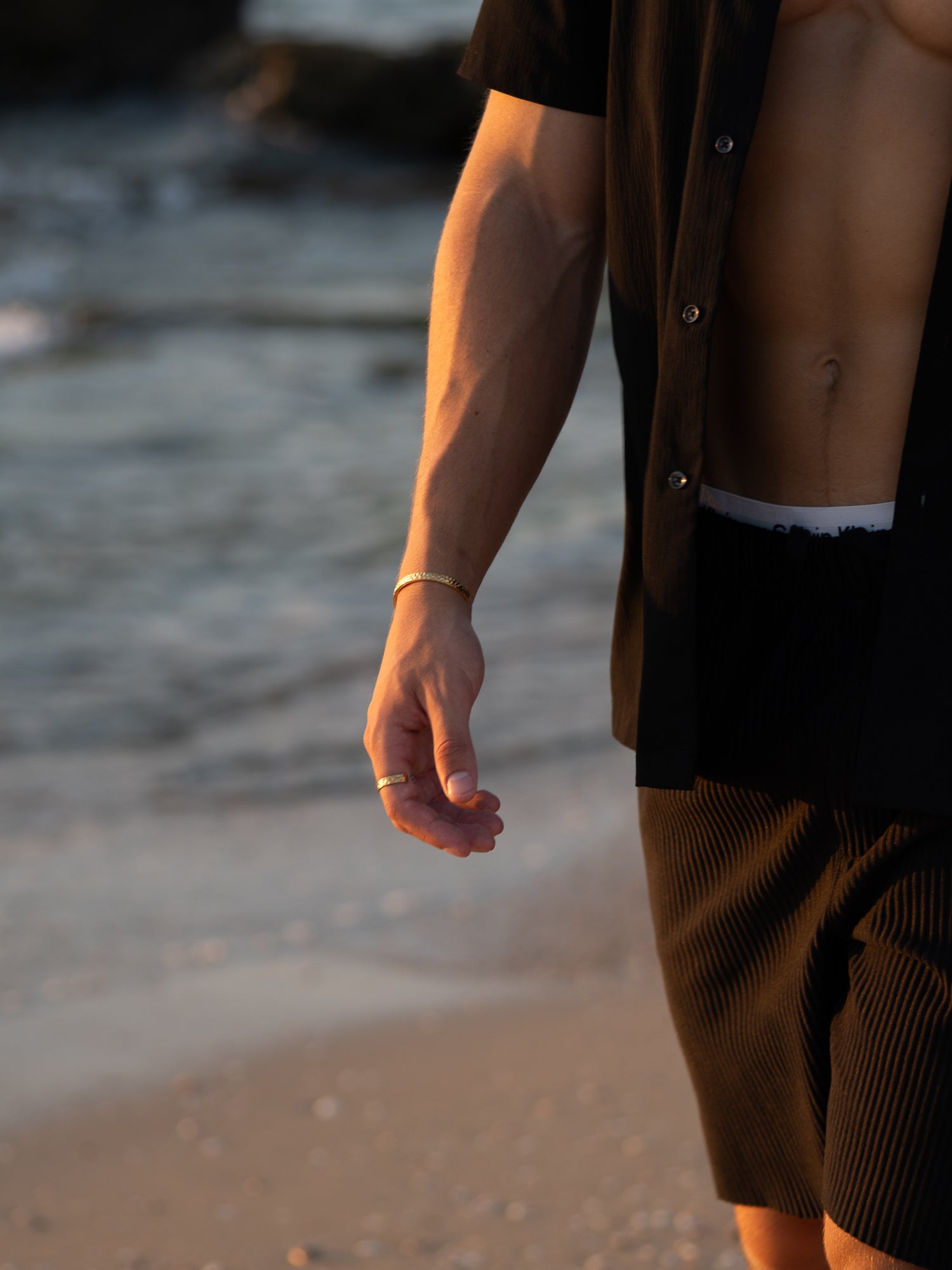 תקריב על יד עונדת צמיד Meduza לגבר בזהב, על רקע חוף ים.