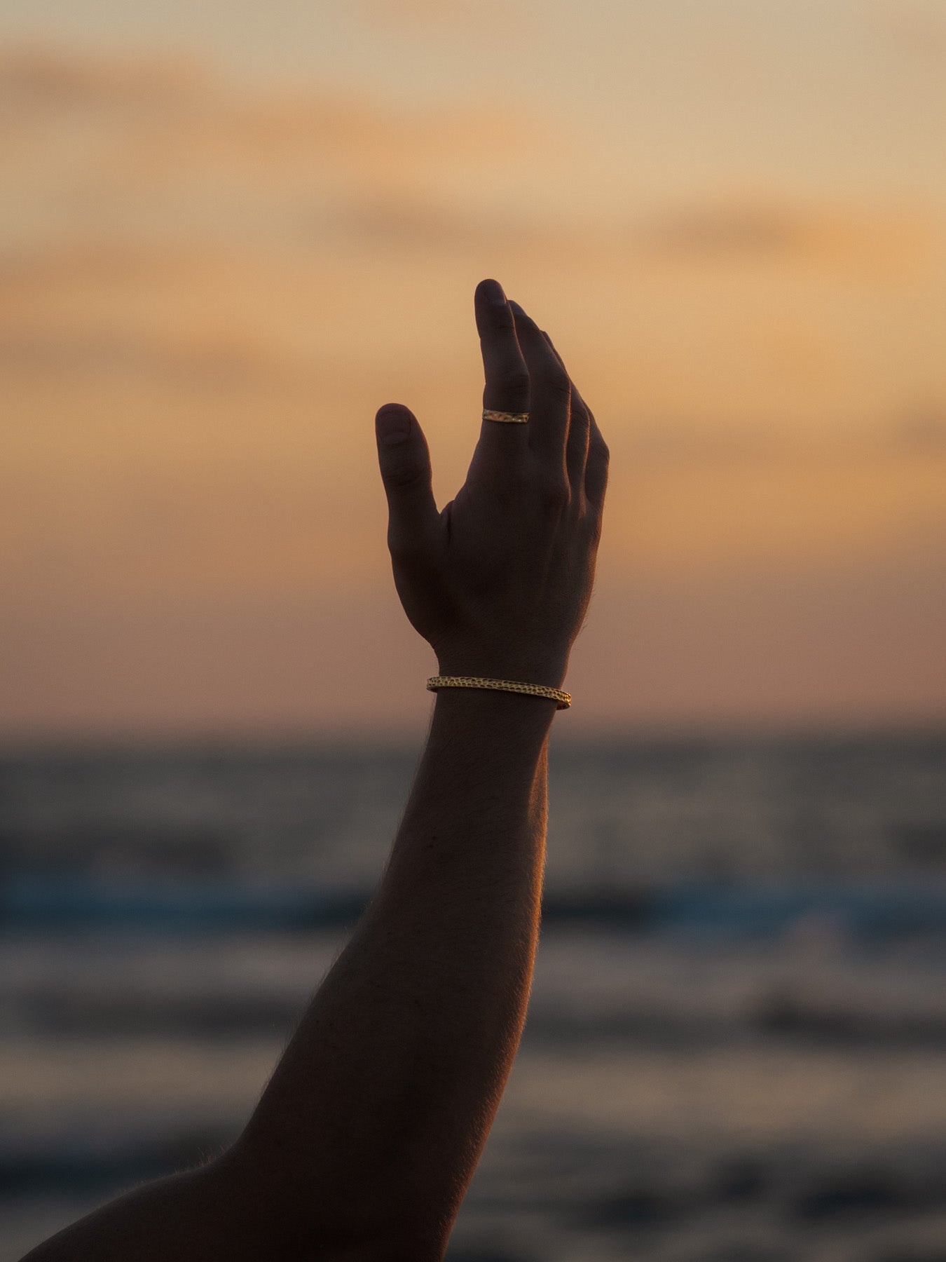 יד גברית מורמת עונדת צמיד וטבעת Meduza בזהב, עם רקע שמיים ושקיעה מעל הים.