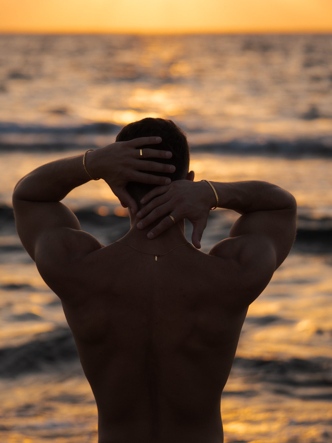 דגם גברי עם גבו לים עונד צמיד וכפת זהב מסוג Meduza ושרשרת NERO, בצילום על רקע שקיעה.