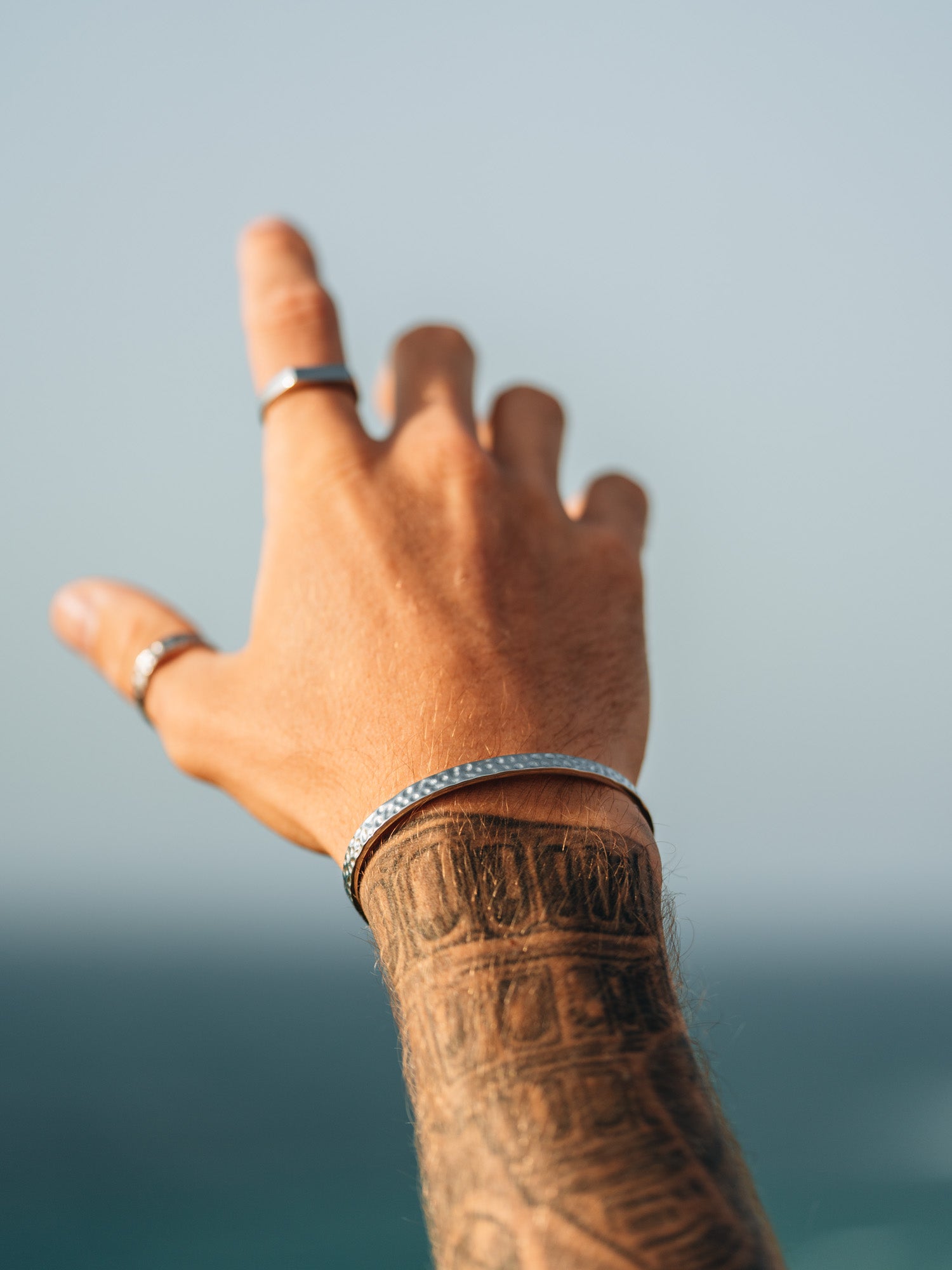 צמיד Meduza כסף על פרק כף יד עם קעקוע, מול רקע שמיים כחולים.