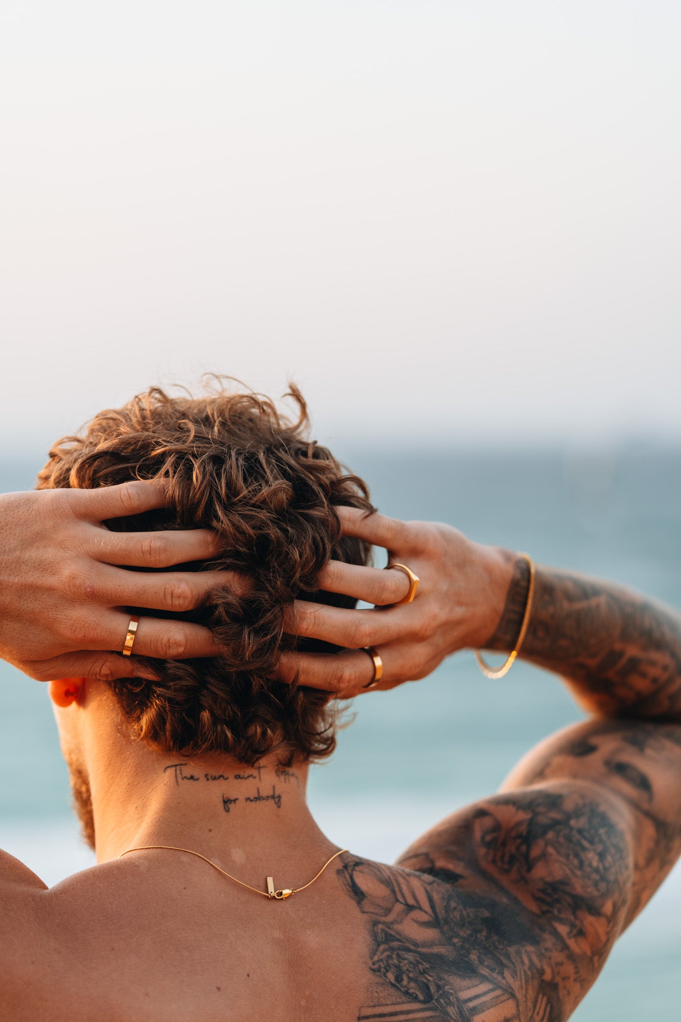 גבר עם שיער מתולתל מצולם מאחור על רקע חוף הים בשקיעה, כשידיו על ראשו ומציגות טבעות זהב, צמידים וקעקועים מפורטים על זרועו וגבו.