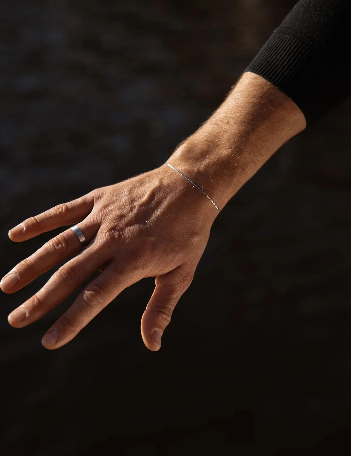 יד של גבר עונדת צמיד Barracuda דק בציפוי כסף בתאורה טבעית, על רקע כהה