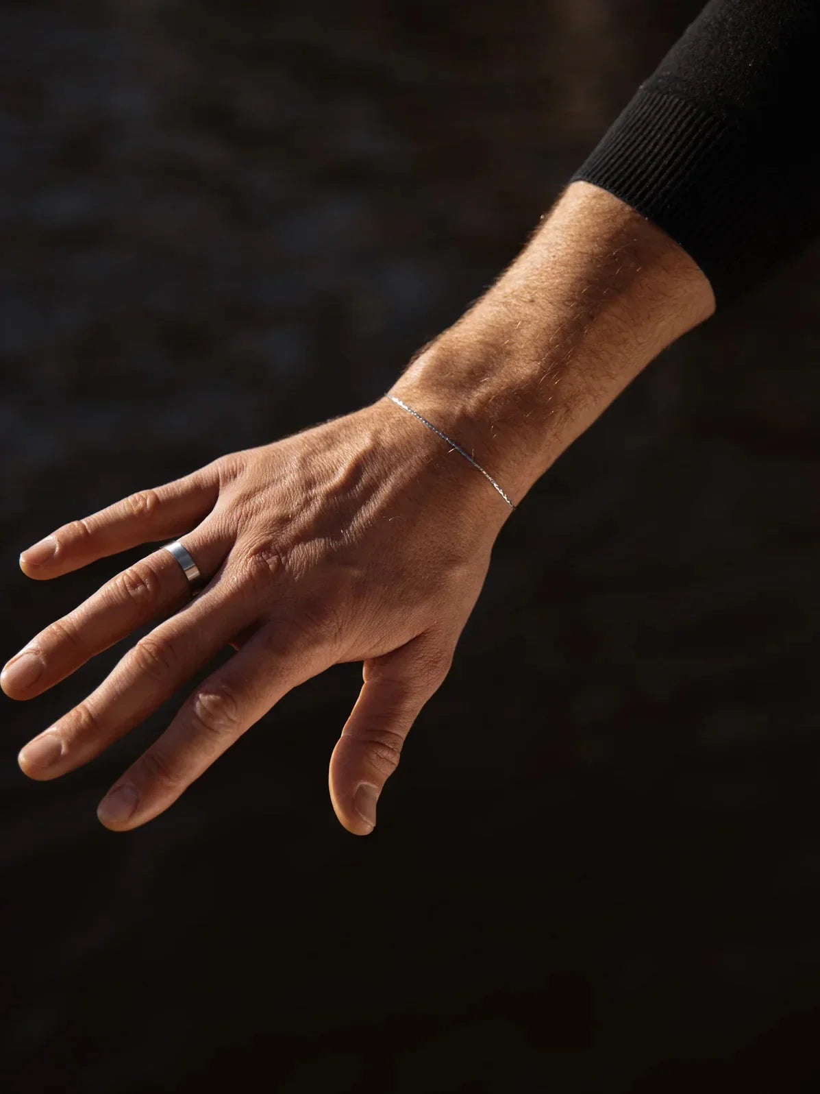 יד של גבר עונדת צמיד Barracuda דק בציפוי כסף בתאורה טבעית, על רקע כהה
