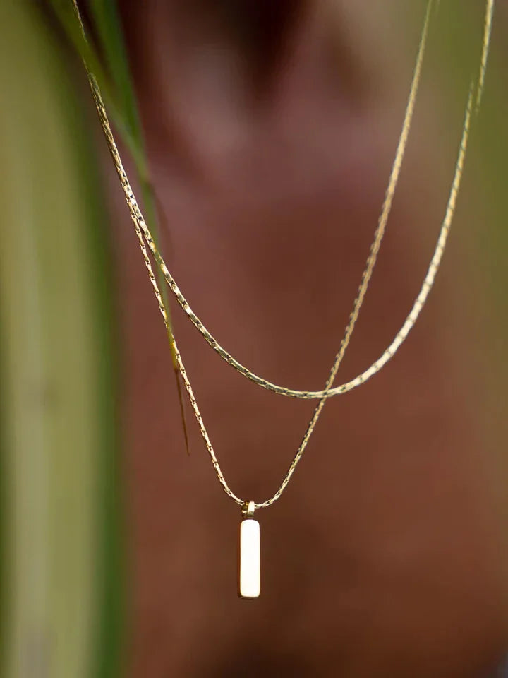 שרשרת זהב מינימליסטית עם תליון NERO על גוף, מצולמת בין עלים באור טבעי