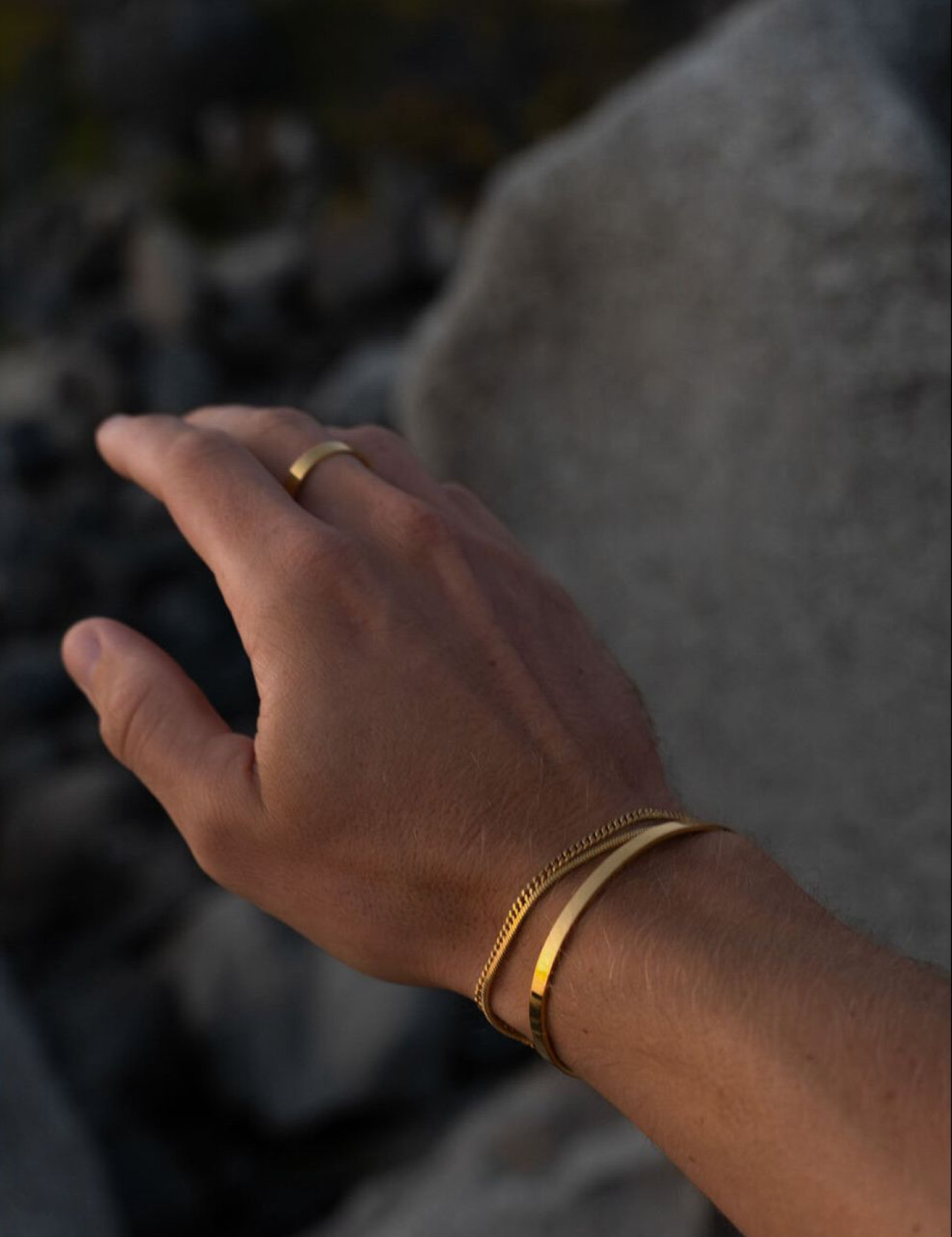 סט תכשיטי זהב לגבר מבית NERO הכולל צמיד קשיח (Cuff) דק ומבריק, צמיד שרשרת גורמט עדינה וטבעת זהב מינימליסטית על רקע אבן טבעית.