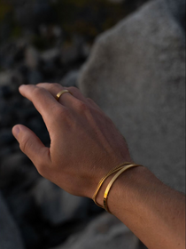 סט תכשיטי זהב לגבר מבית NERO הכולל צמיד קשיח (Cuff) דק ומבריק, צמיד שרשרת גורמט עדינה וטבעת זהב מינימליסטית על רקע אבן טבעית.