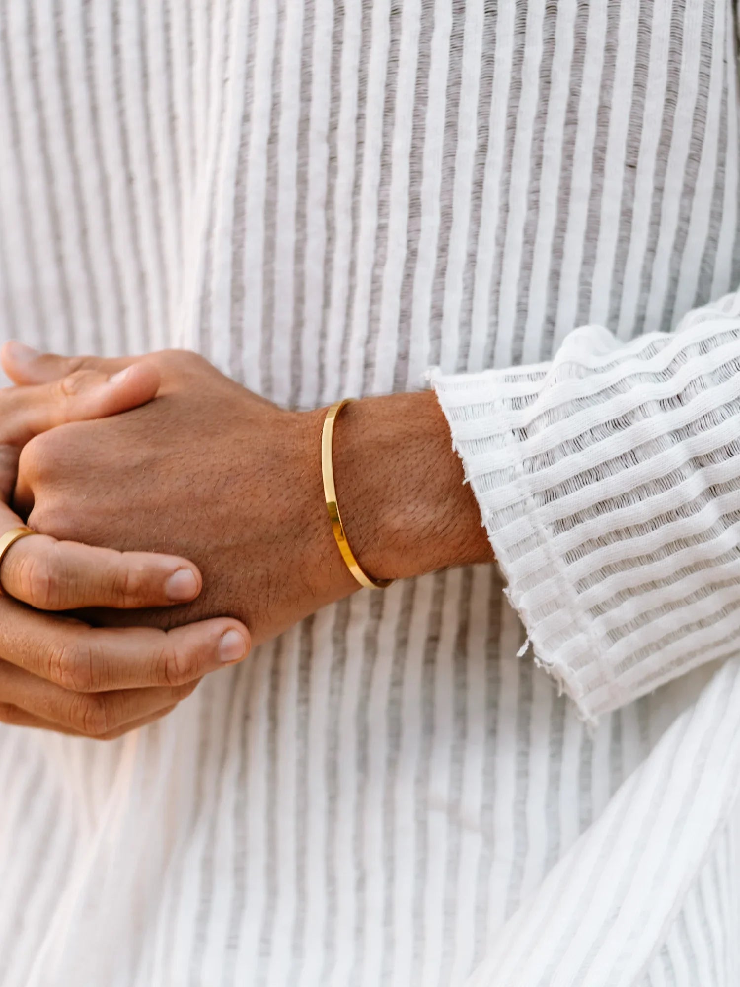 יד של גבר עם חולצה לבנה מפוספסת, לובש צמיד פתוח מוזהב דק בסגנון מינימליסטי.