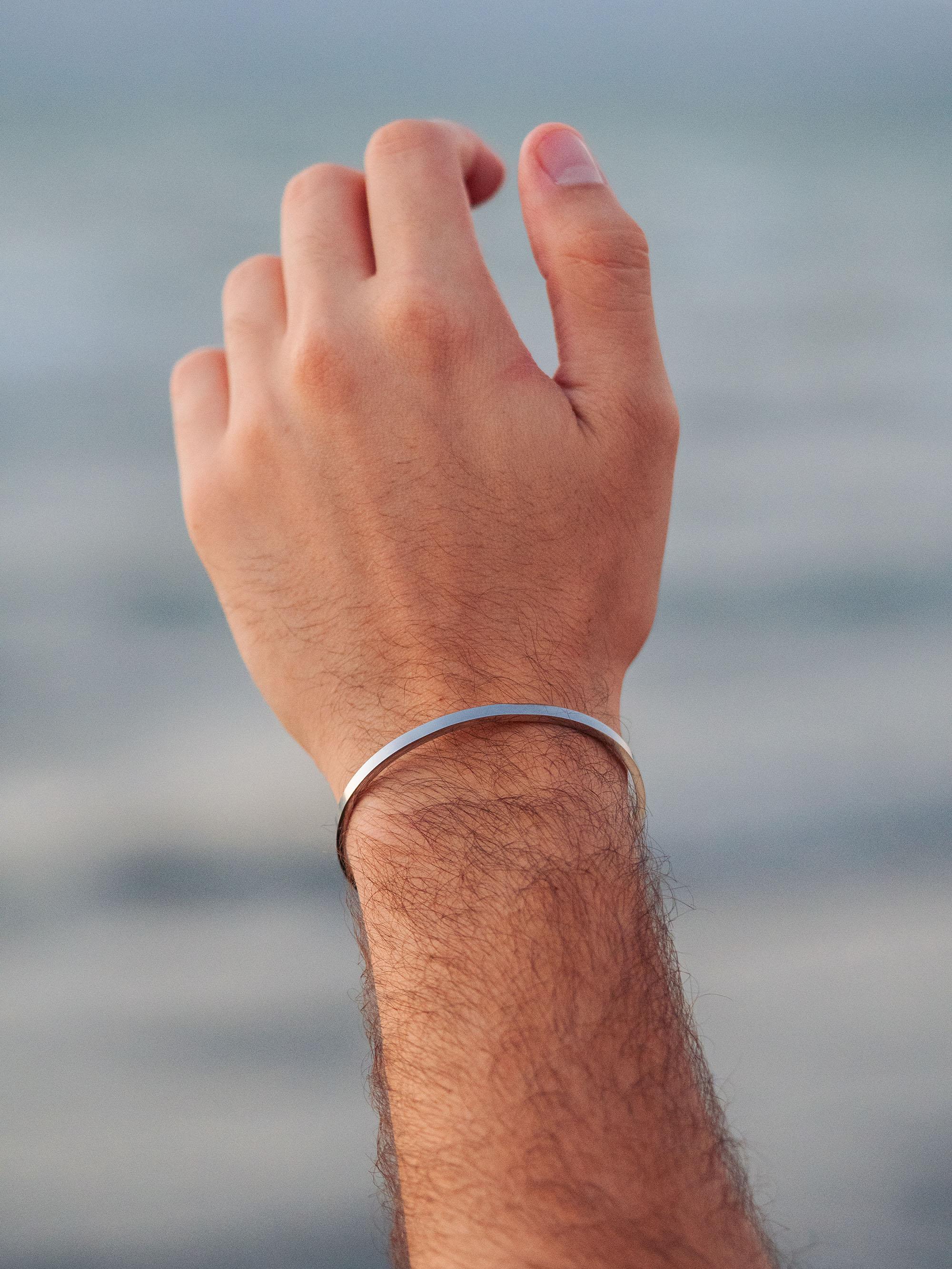 יד של גבר מורמת מול הים, לובש צמיד כסף פתוח ומינימליסטי על פרק כף היד.